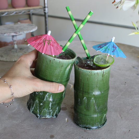 Green ceramic bamboo tumblers with paper bamboo straws and cocktail umbrellas on a gray background